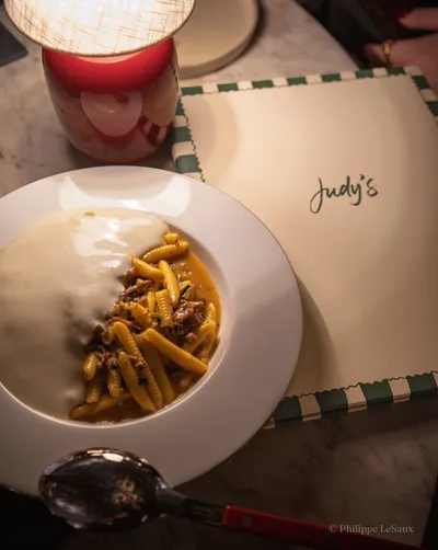 A bowl of cavatelli with braised meat beside a Judy's menu on a marble table