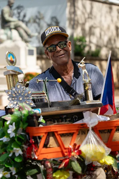 A smiling man surrounded by trophies, flowers, and a Puerto Rican flag