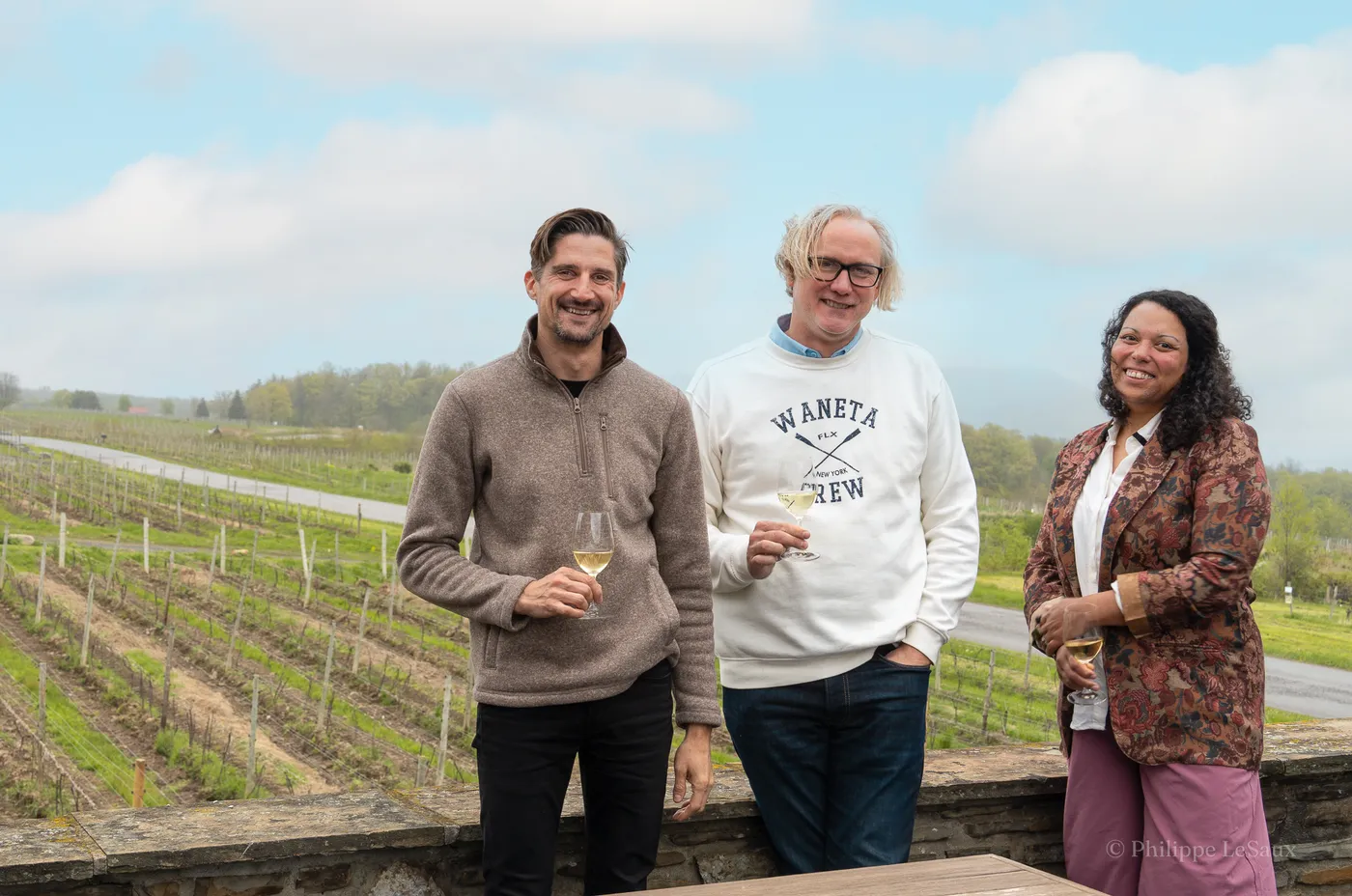 Three people with wine glasses overlooking rows of grapevines at a vineyard
