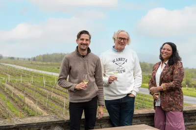 Three people with wine glasses overlooking rows of grapevines at a vineyard